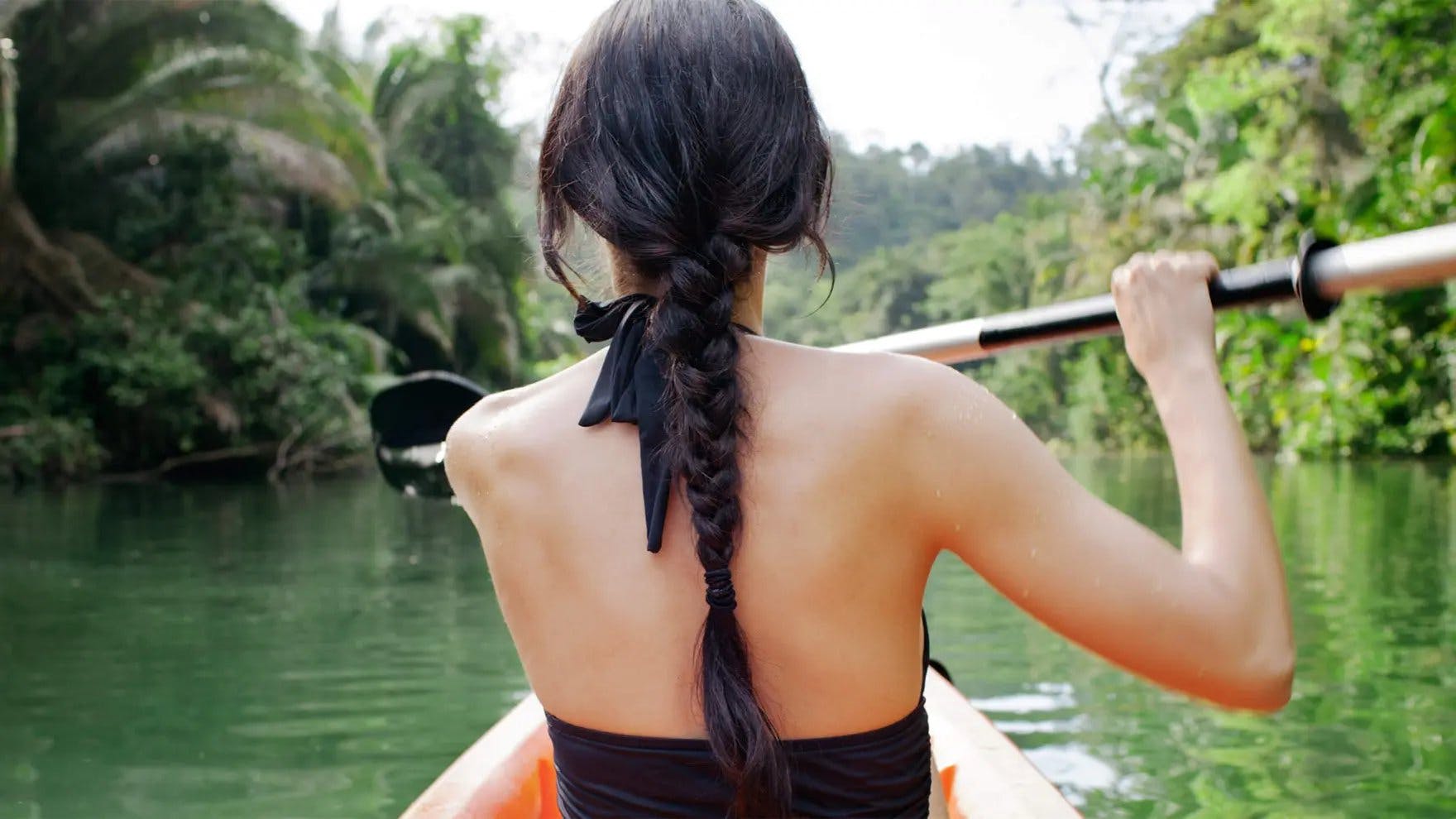 Woman with long, black hair in a braid kayaking down a river with her back turned to the camera
